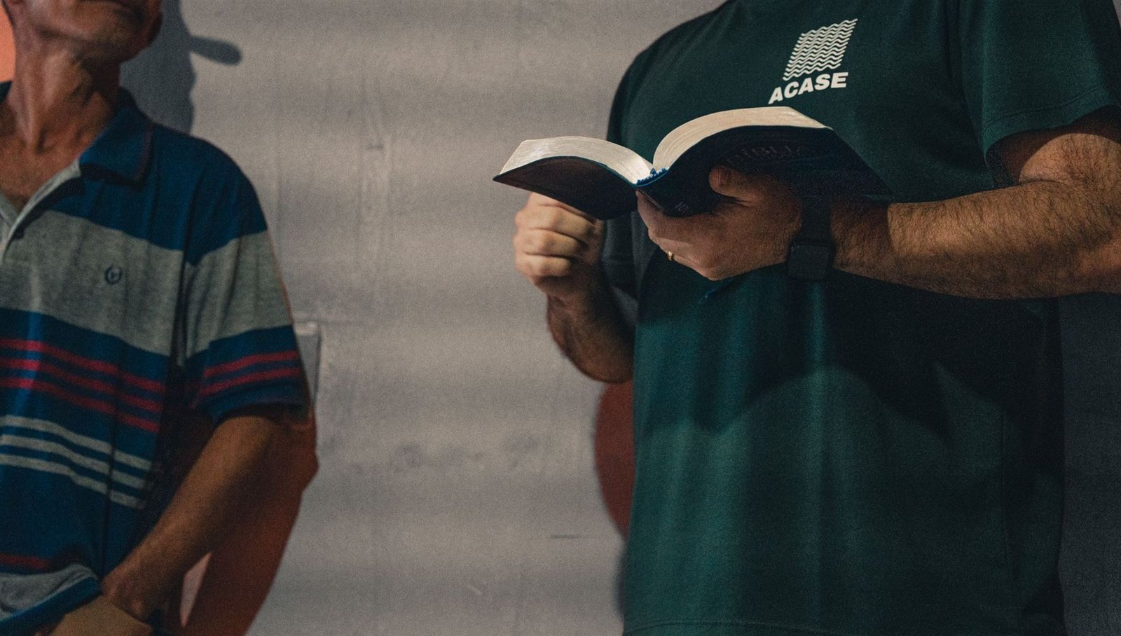 Voluntário com a camiseta da ACASE lê a Bíblia ao lado de um senhor.