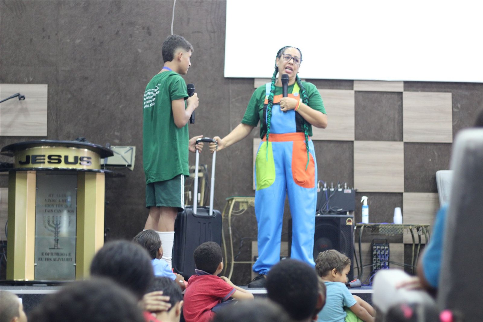 Voluntária da ACASE fala ao microfone para crianças sentadas diante do palco, ao lado de um voluntário adolescente.